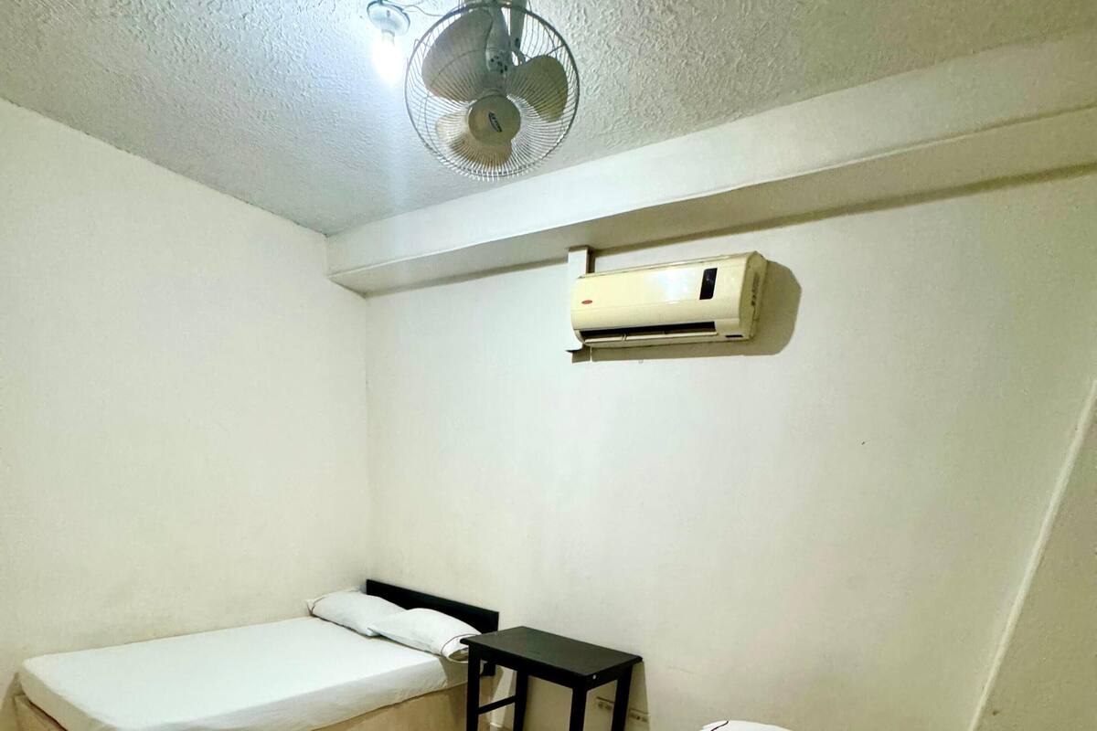 A room with two single beds, a small table, a ceiling fan, and an air conditioning unit.
