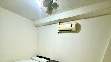 A room with two single beds, a small table, a ceiling fan, and an air conditioning unit.