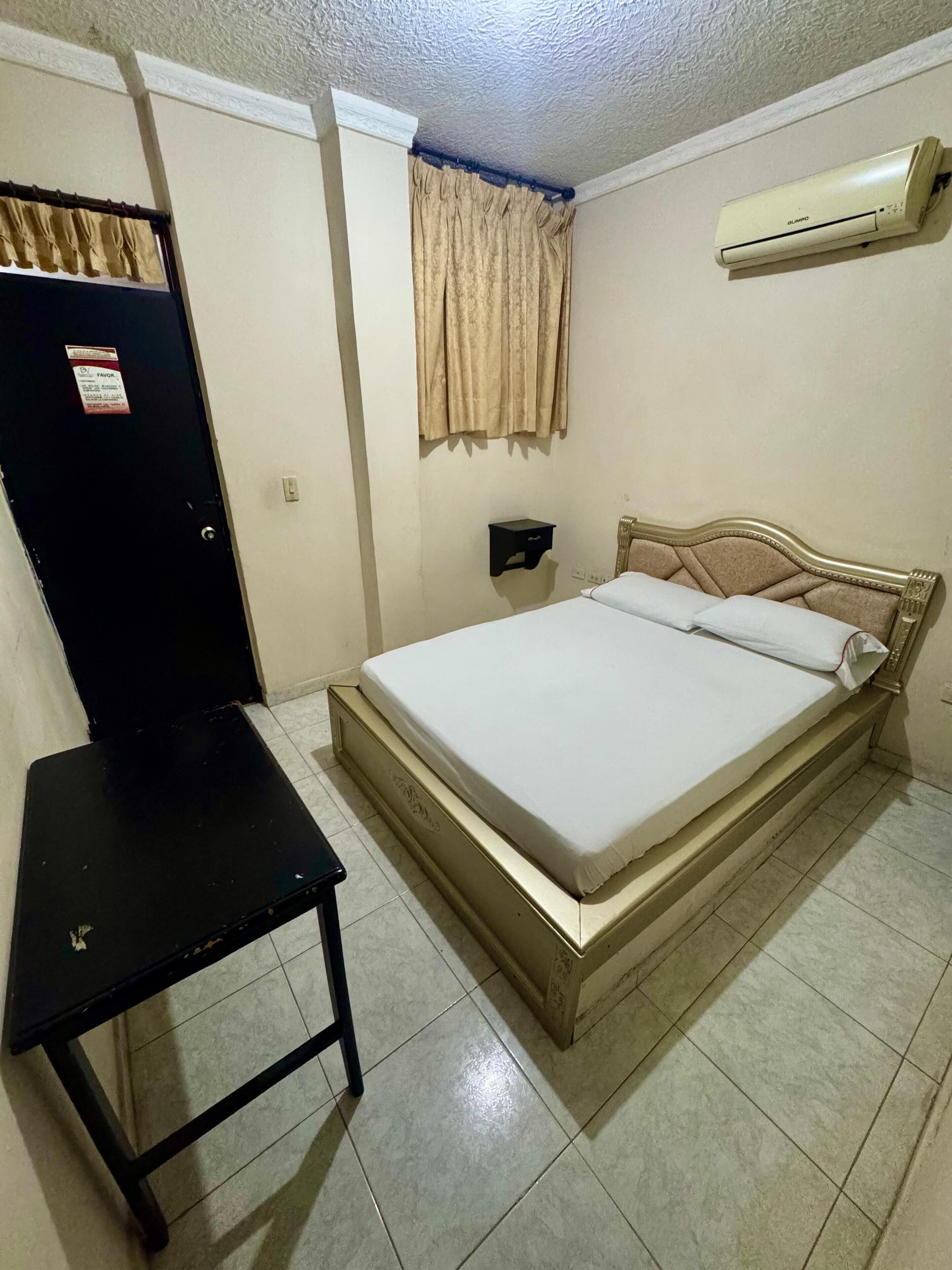 A bedroom with a bed, a small table, a wall-mounted air conditioner, and a door with a sign.