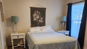 Un dormitorio con una cama, dos mesitas de noche, un ventilador de techo, una obra de arte en la pared y una ventana con cortinas.