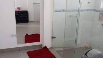 A bathroom with a glass shower enclosure, a red rug, and a grey trash bin.