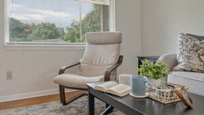 Un salon moderne avec un fauteuil à revêtement gris, une table basse sur laquelle se trouvent un livre et une tasse bleue, et une fenêtre avec des stores.