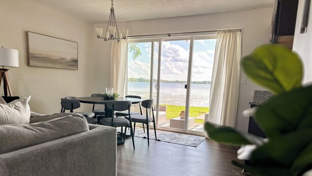 Un salon moderne avec un canapé gris, une table à manger avec des chaises, un lustre et une vue sur un lac, visible à travers de grandes portes coulissantes.