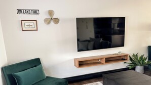 Un salon avec un canapé vert, un téléviseur à écran plat fixé au mur, une étagère en bois située sous le téléviseur, une plante en pot et une table basse.