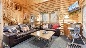 A log cabin interior with a wooden staircase, leather sofas, a coffee table, and a television mounted on the wall.