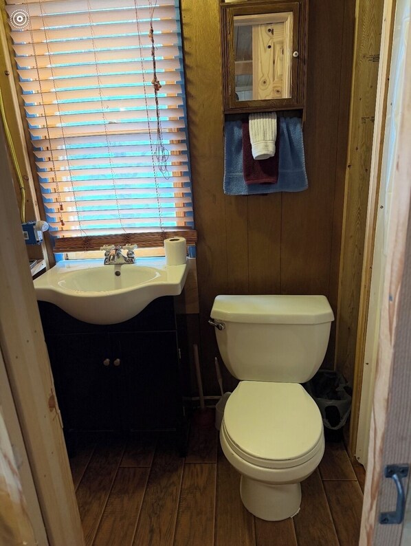 Un baño pequeño con suelo de madera, un lavamanos blanco, un inodoro y una ventana con persianas.