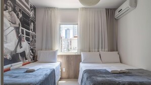 Ein Hotelzimmer mit zwei Betten, einem großen Fenster mit Vorhängen, einer fest installierten Klimaanlage und einem Wandbild.