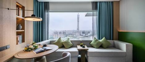 A modern hotel room with a city view, a sofa, a dining table, and a bookshelf.
