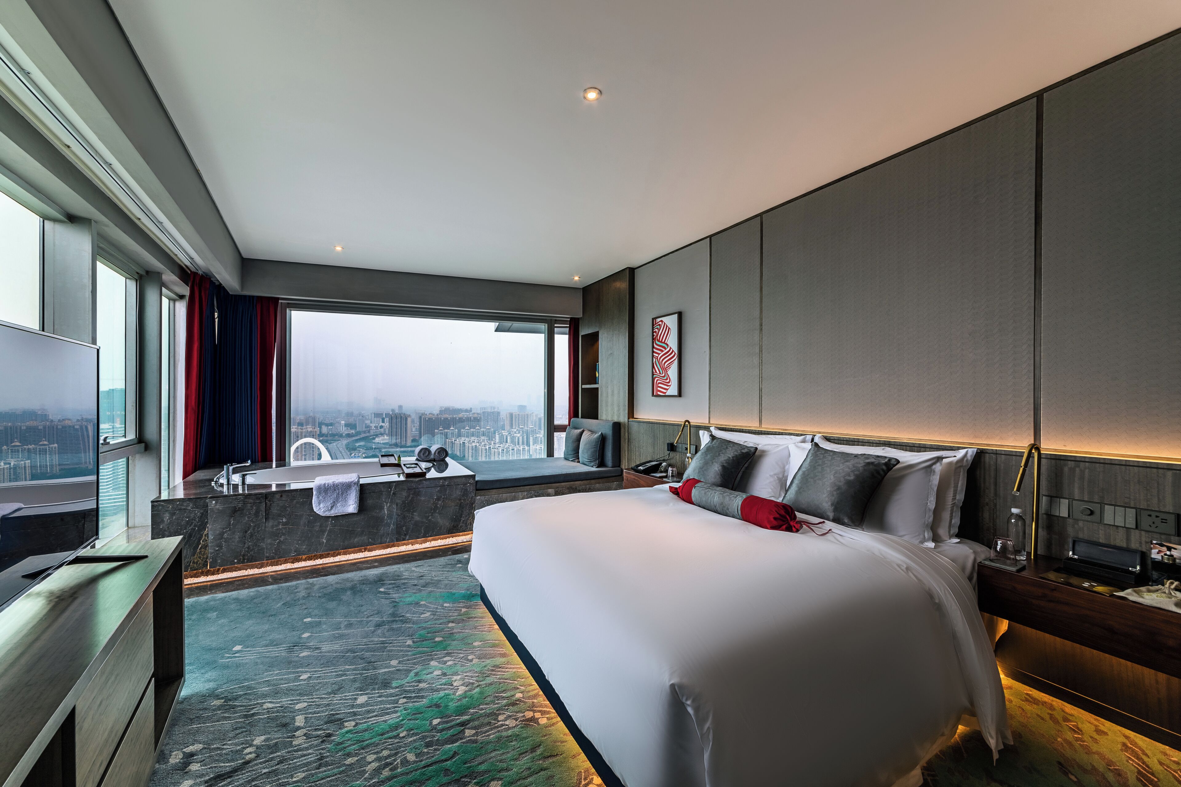 A modern hotel room with a large bed, a city view, a bathroom with a bathtub, and a desk with a computer.