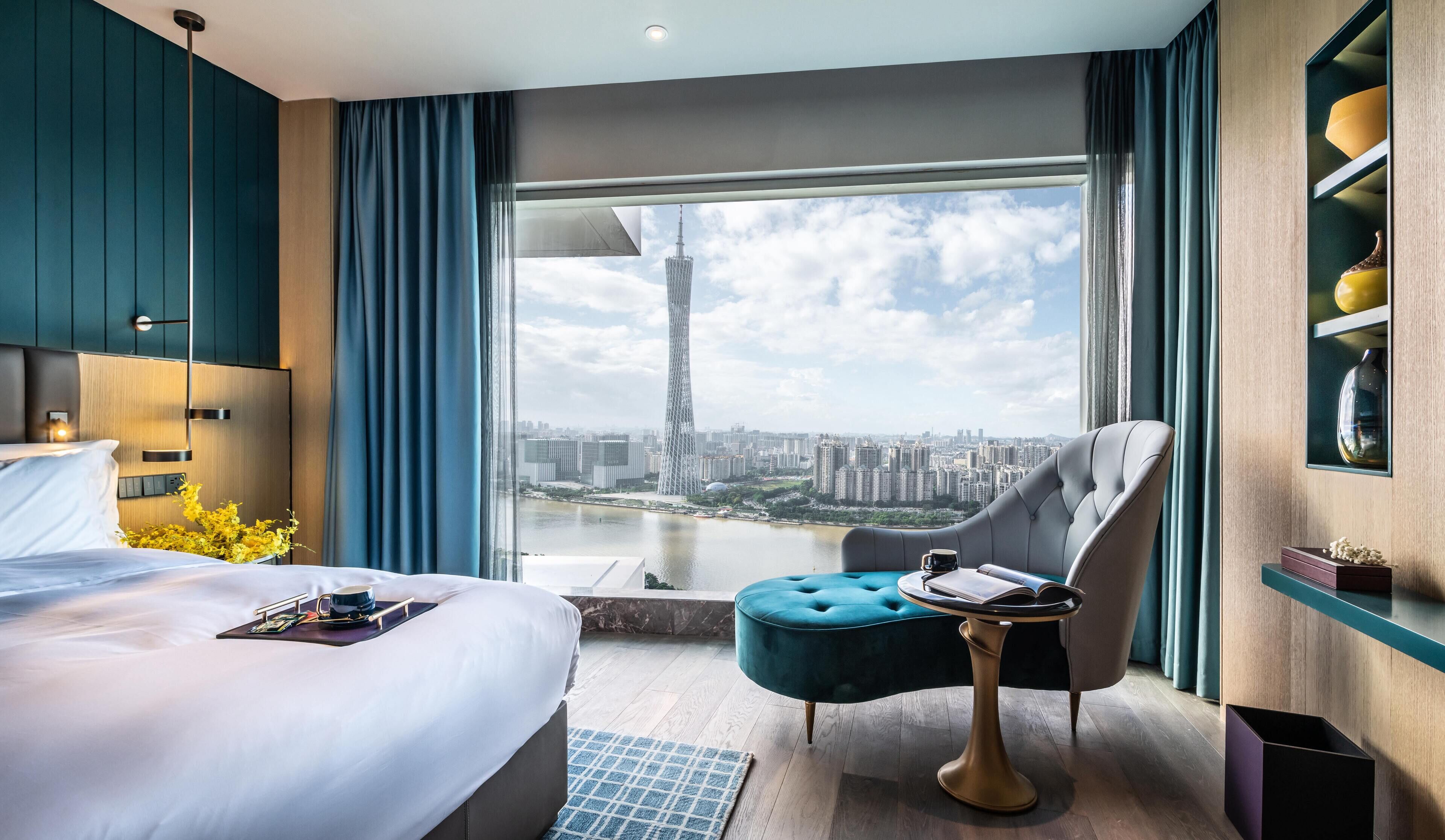 A modern hotel room with a large bed, a tufted armchair, a small round table, and a view of a cityscape and a tall tower through a large window.