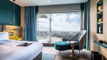 Una habitación de hotel moderna con una cama grande, un sillón capitonado, una pequeña mesa redonda y vistas a un paisaje urbano y una torre alta a través de un amplio ventanal.
