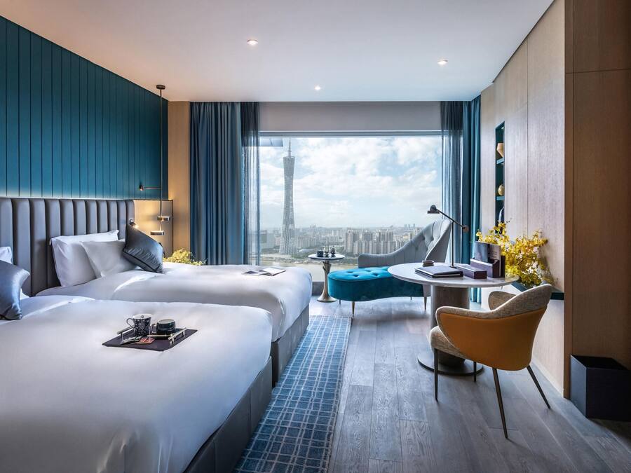 A modern hotel room with two beds, a seating area, and a large window with a city view.