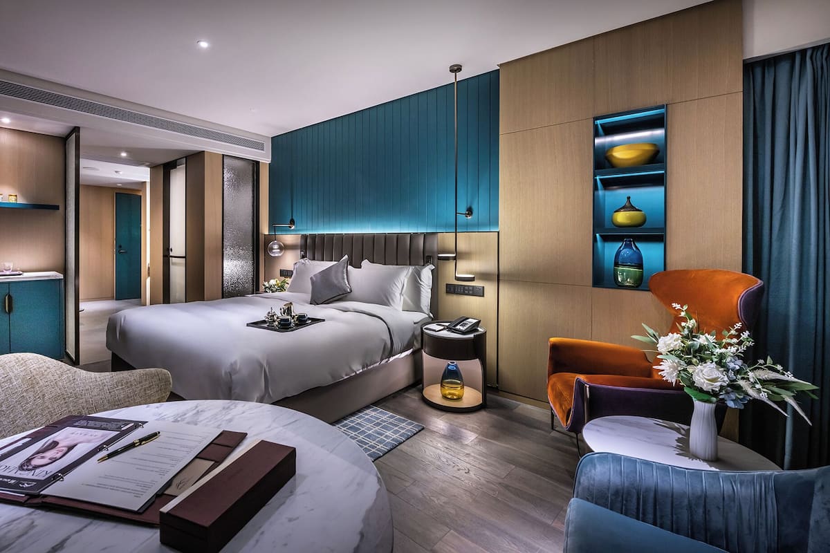 Una habitación de hotel moderna con una cama grande, un escritorio con una lámpara, una silla, una mesita con un florero y un minibar.