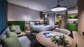 A modern hotel room with a bed, a sofa, a round coffee table with a magazine and flowers, a small side table, and a view into the bathroom and kitchen area.