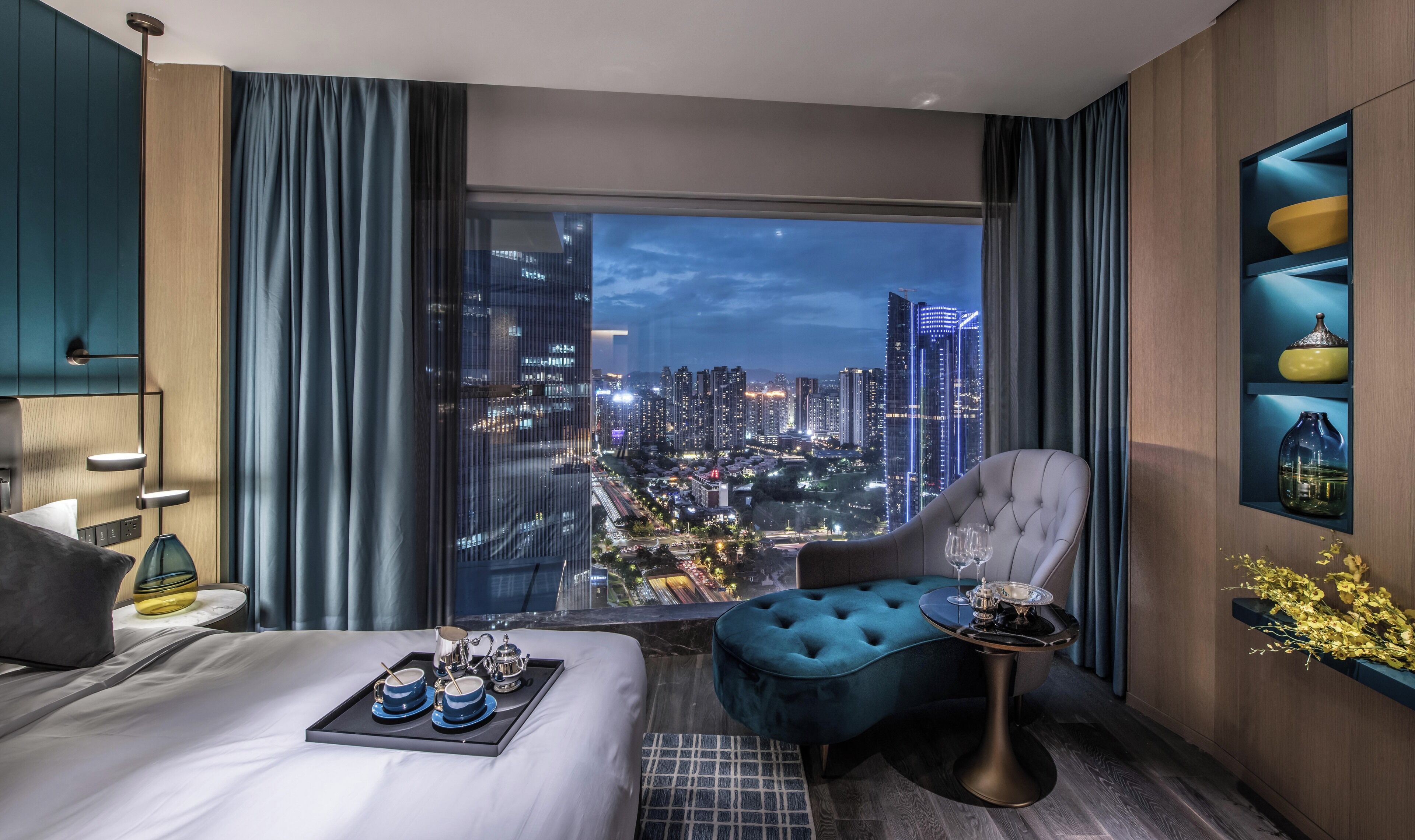 Una habitación de hotel moderna con un ventanal grande que ofrece vistas a la ciudad, una cama cómoda, un área de descanso y una mesita con una bandeja de té y tazas.