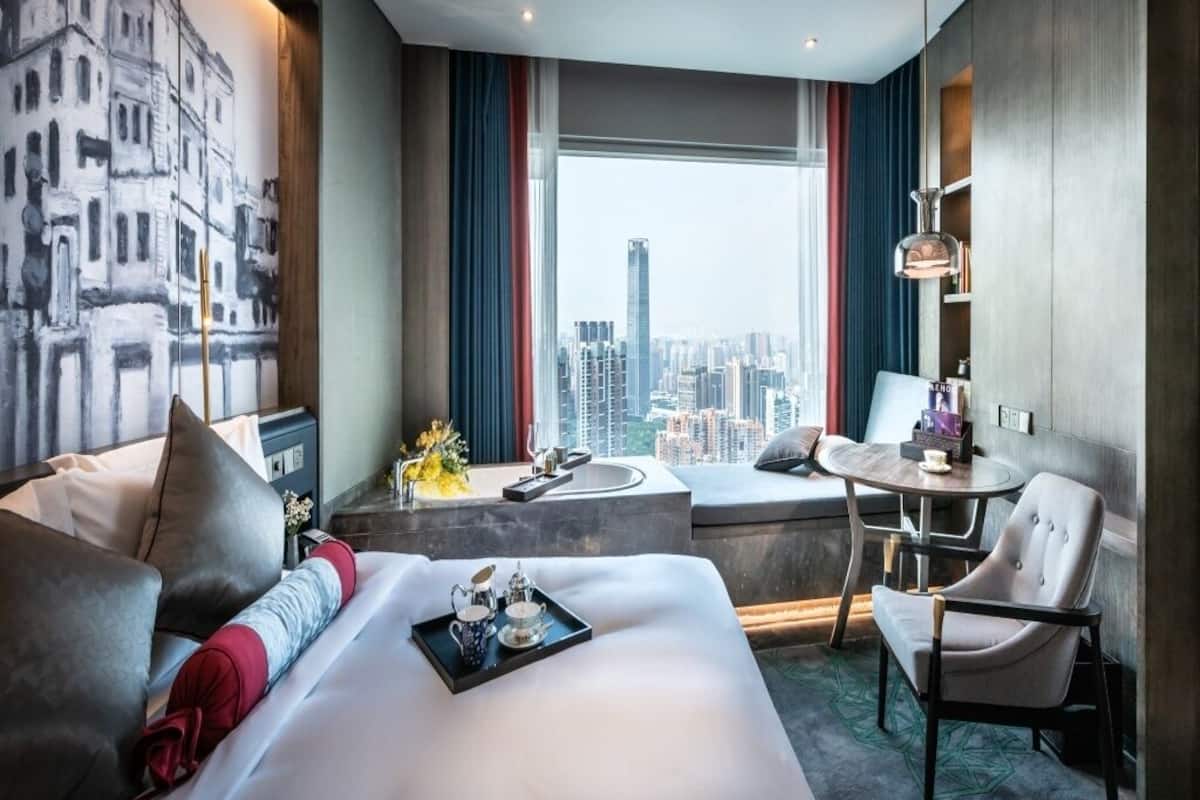 Habitación de hotel con una cama grande, zona de estar con una mesa y sillas, vista del paisaje urbano a través de un ventanal amplio y un cuadro en la pared.