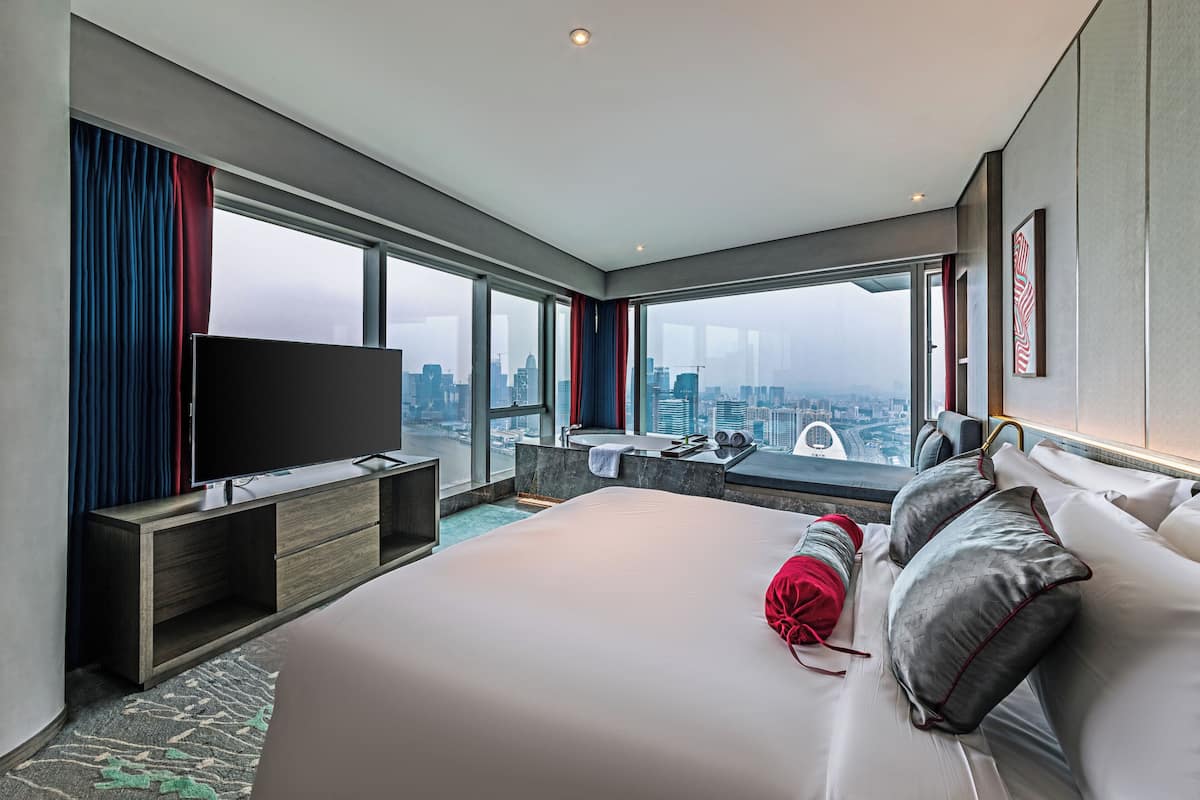 Habitación de hotel moderna con una cama grande, televisor de pantalla plana, vista a la ciudad, un escritorio pequeño con lavamanos y una ventana con cortinas rojas.