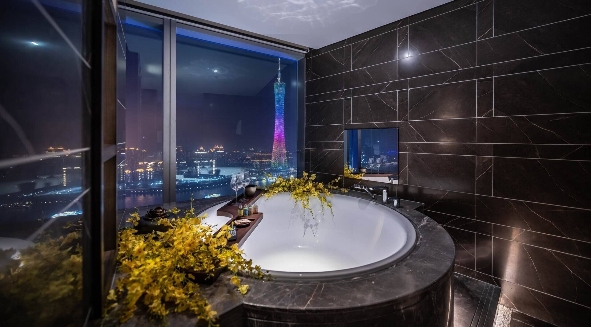 A modern bathroom with a large bathtub, a cityscape view, and a television on the wall.