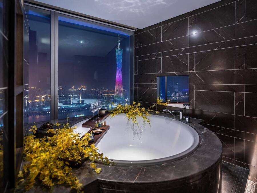 A modern bathroom with a large bathtub, a cityscape view, and a television on the wall.