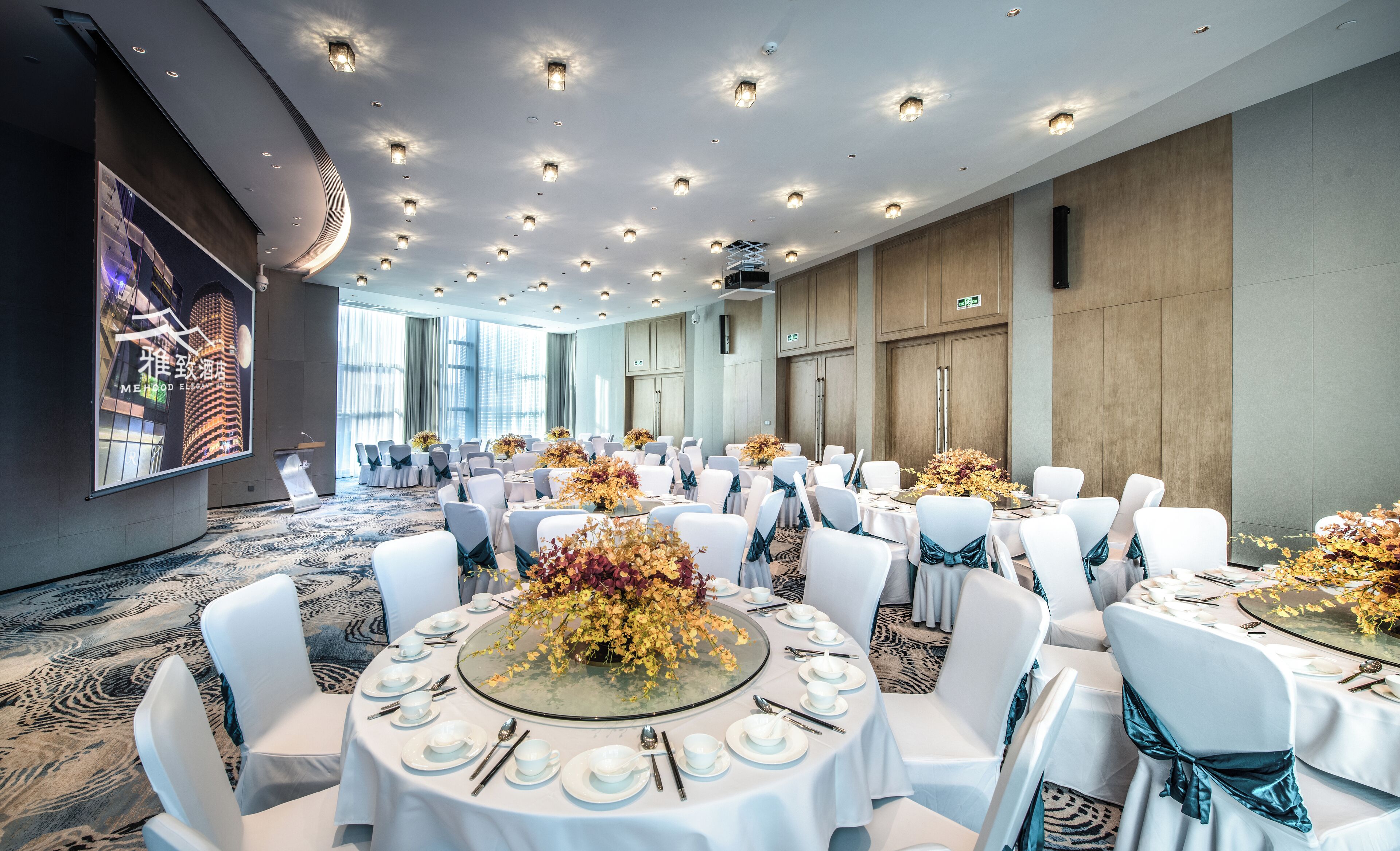 A banquet hall with round tables set for a formal event, featuring white tablecloths, chairs with blue sashes, and floral centerpieces.