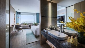 Een moderne hotelkamer met een groot bed, een zithoek met een bank en fauteuil, een ruime badkamer met wastafel en spiegel, en uitzicht op de stad.