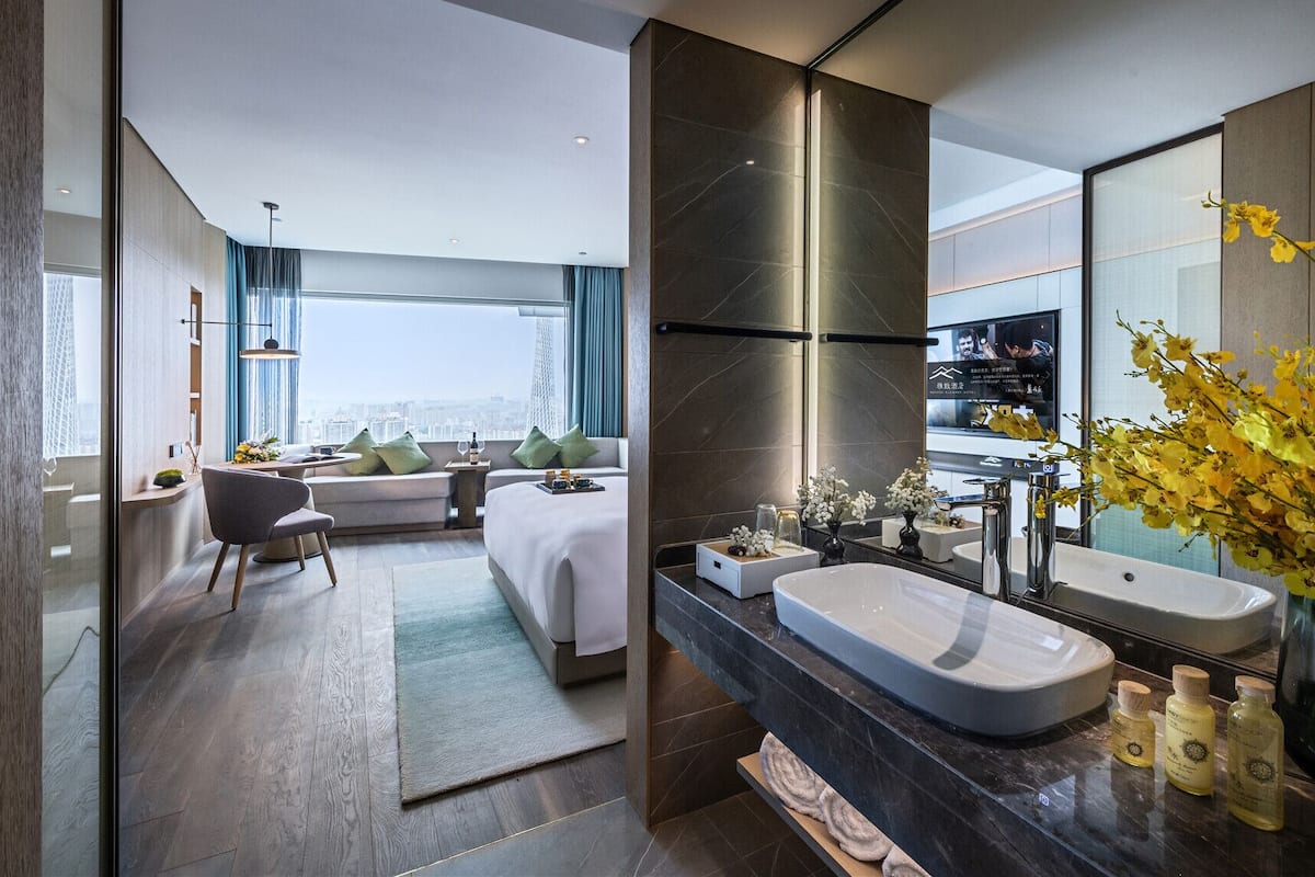 Una habitación de hotel moderna con una cama grande, una zona de estar con sofá y sillón, un baño amplio con tocador y espejo, y vistas a la ciudad.
