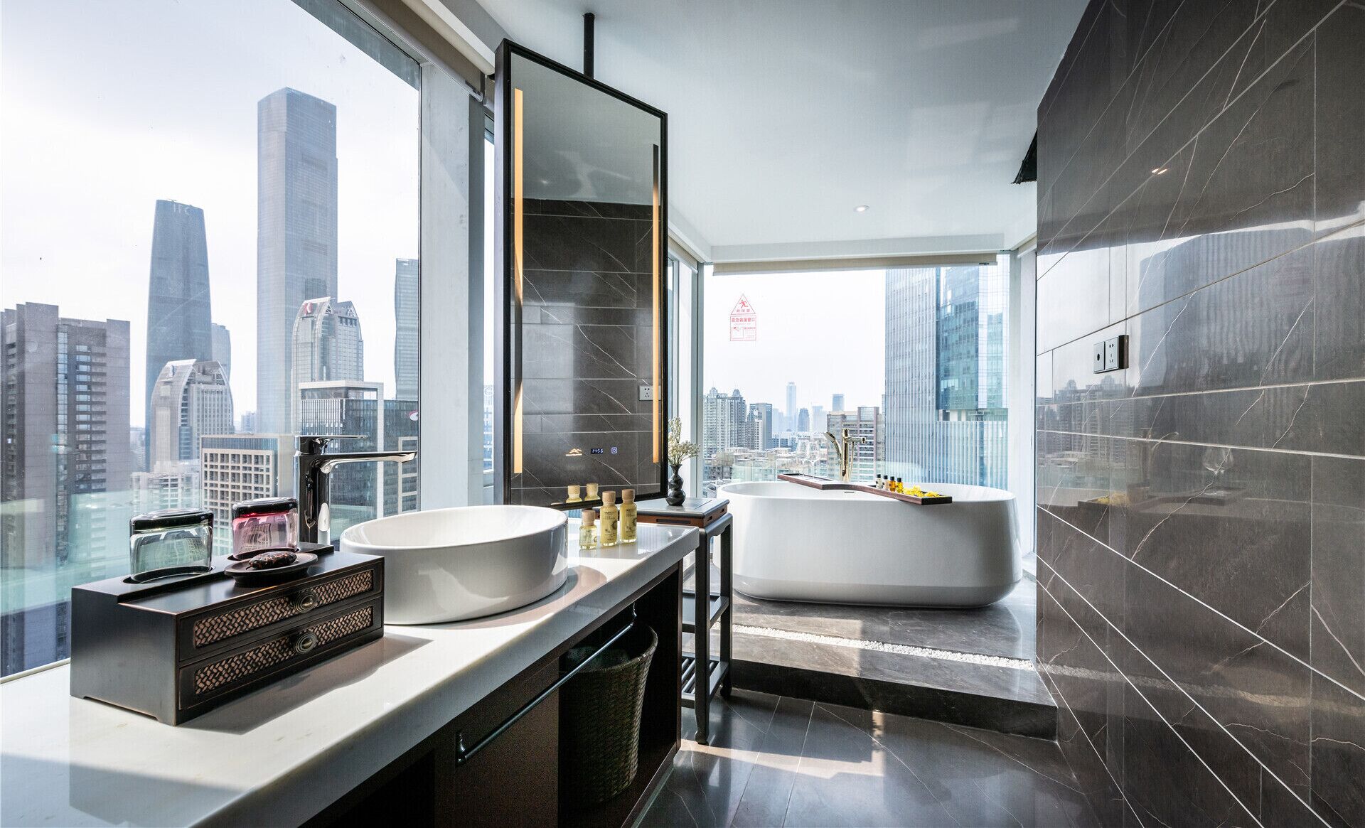 A modern bathroom with a large sink, a bathtub, and a vanity with a mirror. The bathroom has a city view through large windows.