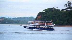 Um barco de vários andares com passageiros, navegando em águas calmas com paisagem costeira ao fundo.