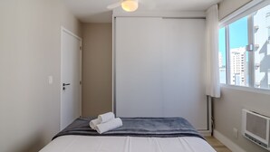 Une chambre à coucher avec un lit, une porte, une fenêtre donnant sur des immeubles et un ventilateur de plafond.