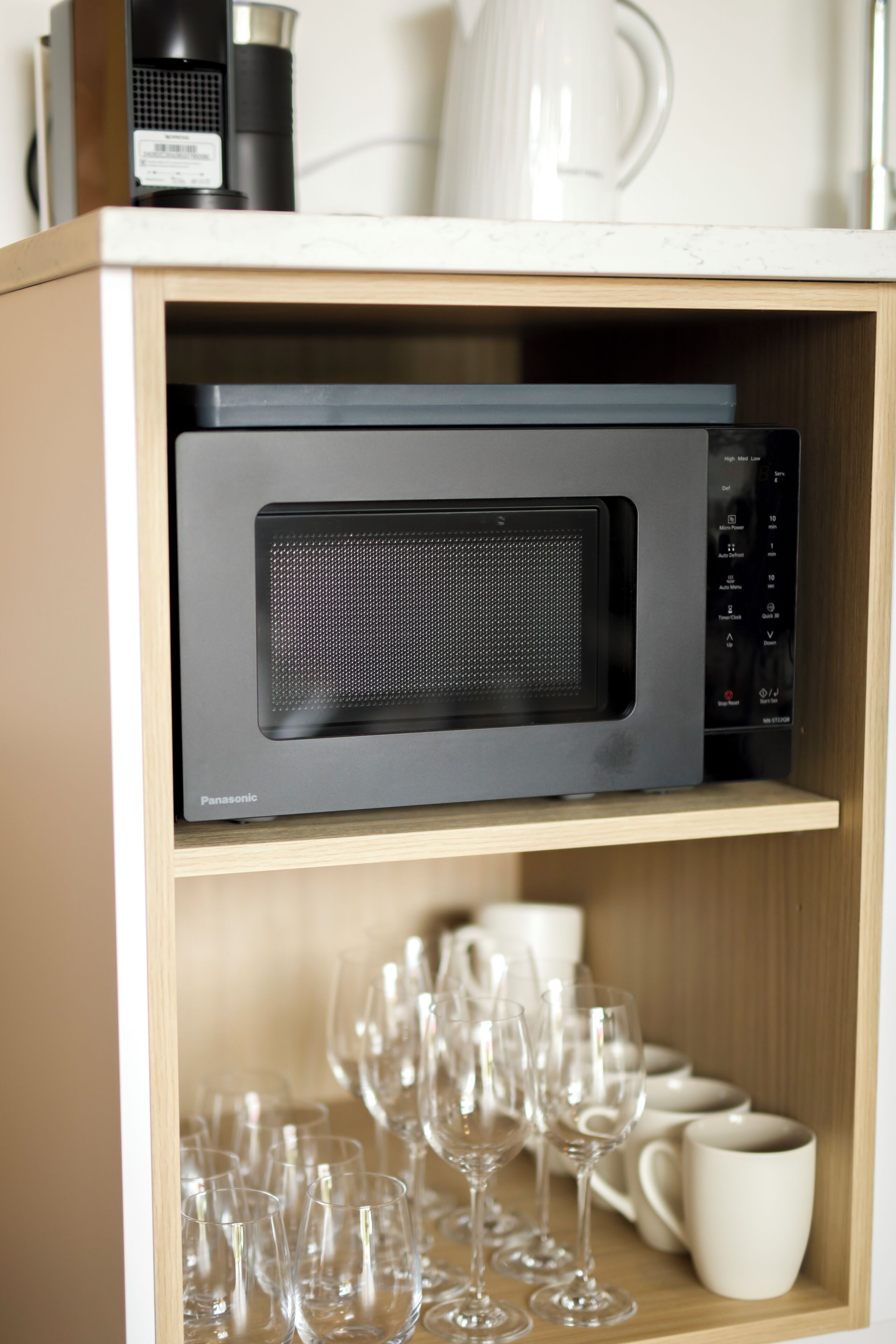 A microwave oven with a control panel, a coffee maker, and a set of wine glasses and cups on a shelf.