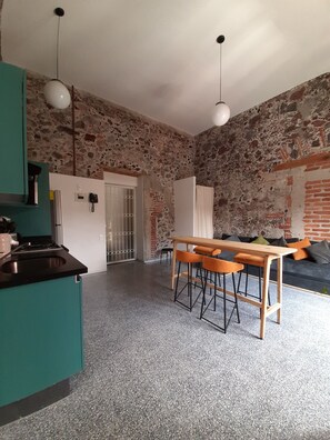 A kitchen with a teal countertop, a dining area with a wooden table and orange chairs, and a stone wall.