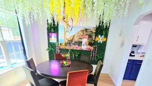 A dining area with a round table and chairs, a kitchen area with blue cabinets, and a wall with a scenic view and greenery.