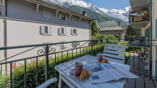 A balcony with a table set for breakfast, a mountain view, and a building with multiple windows.