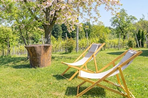 Two wooden lounge chairs with white cushions on a grassy lawn, a large tree with pink blossoms, and a wooden post in the background.