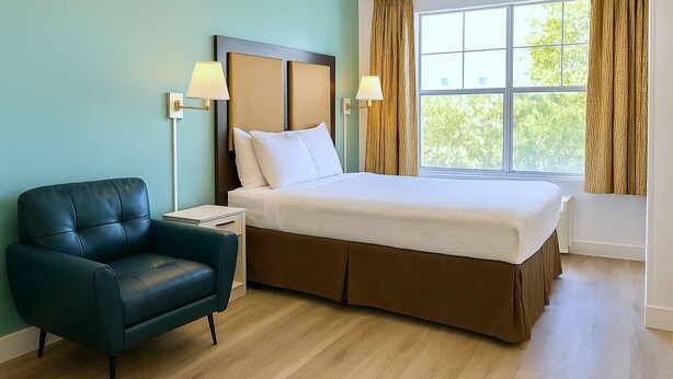 A hotel room with a bed, a nightstand, a chair, and a window with curtains. - Fully Furnished Hotel Suites in Las Vegas with Fitness Room and Outdoor Pool (Las Vegas)
