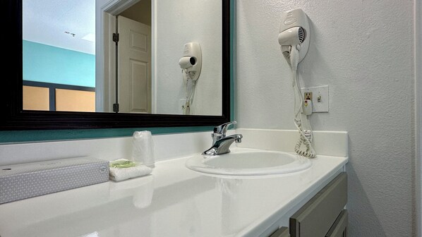 Un baño con un lavamanos blanco, un espejo, un secador de pelo montado en la pared y un dispensador de toallas.
