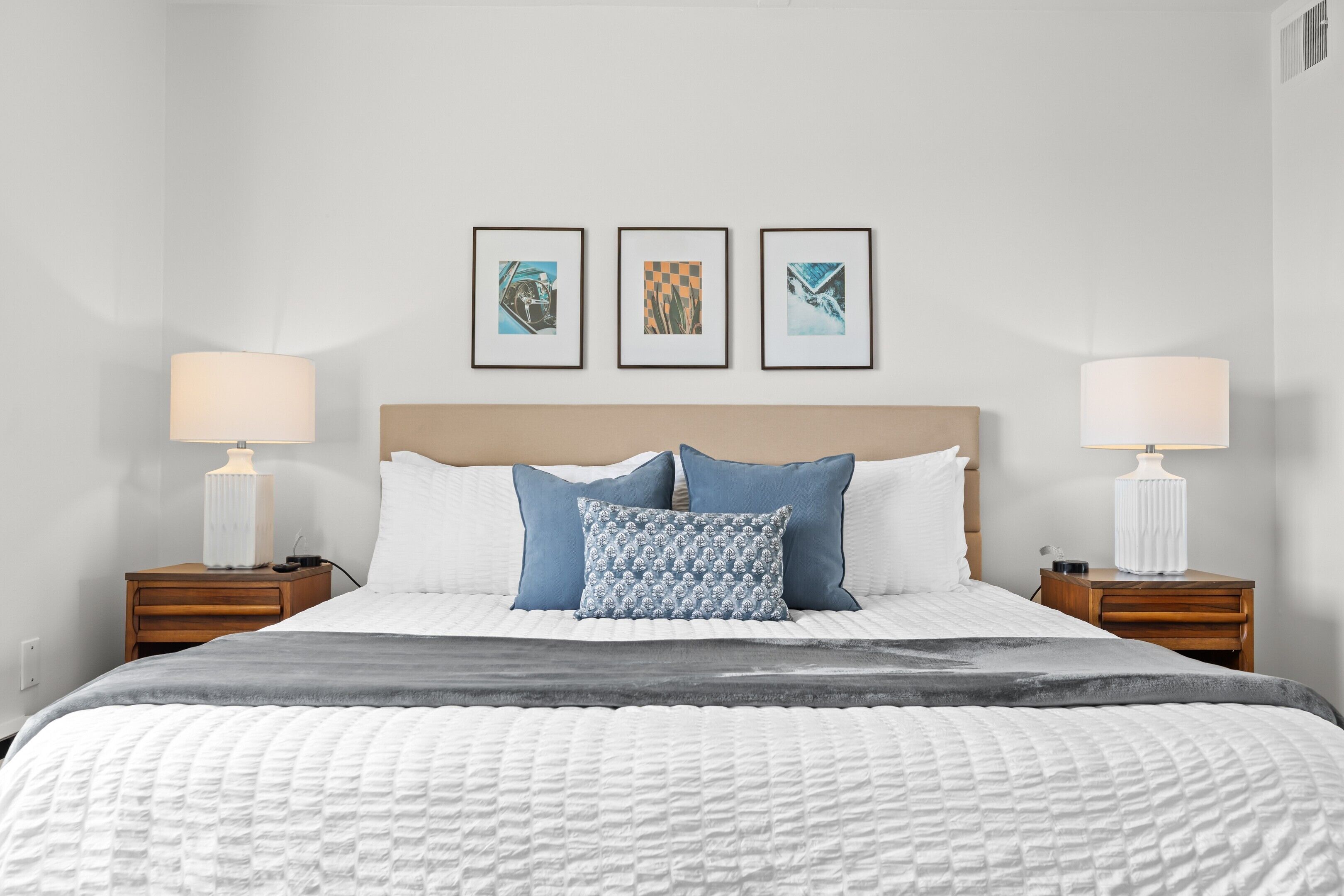 A neatly made bed with a white quilt, blue and white patterned pillows, and a gray throw blanket. There are two bedside tables with lamps, and three framed pictures on the wall above the bed.