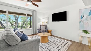 Una sala de estar moderna con un sofá gris, una mesa de centro redonda, un ventilador de techo, un ventanal con vistas a árboles y un televisor montado en la pared.