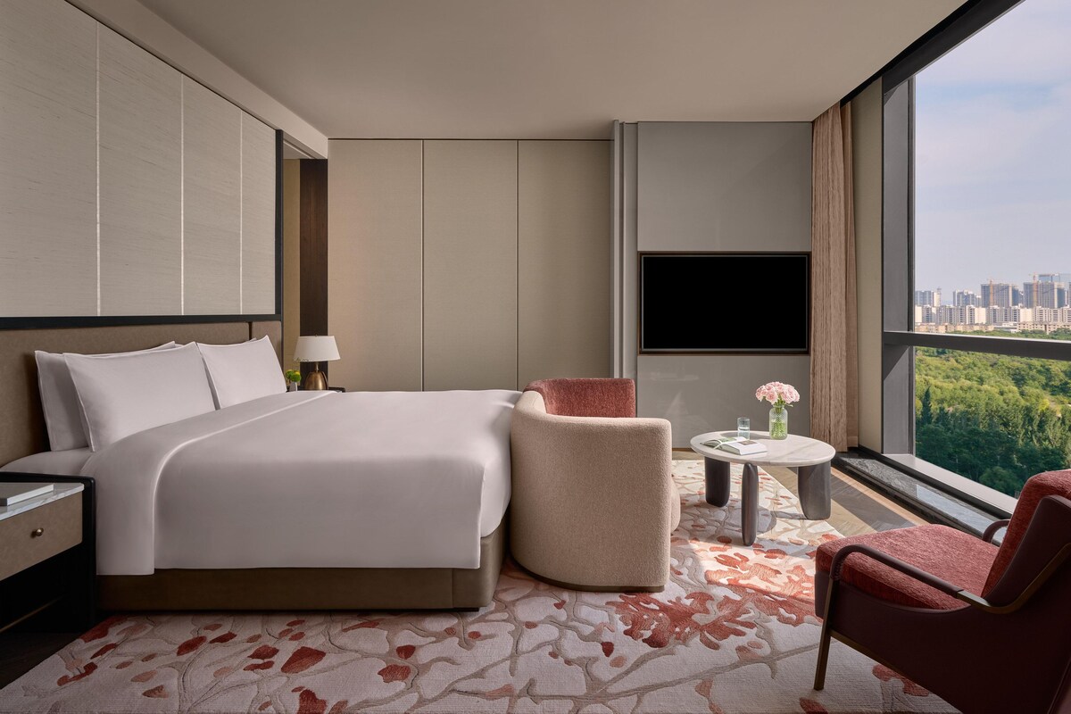 A modern hotel room with a large bed, a seating area with a chair and a small table, a TV mounted on the wall, and a view of a cityscape through large windows.