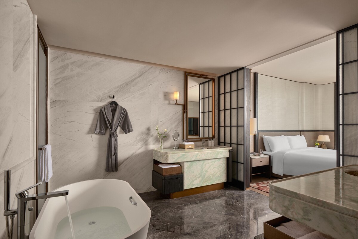 A modern bathroom with a freestanding bathtub, a vanity with a marble countertop, a large mirror, and a bed with white linens.