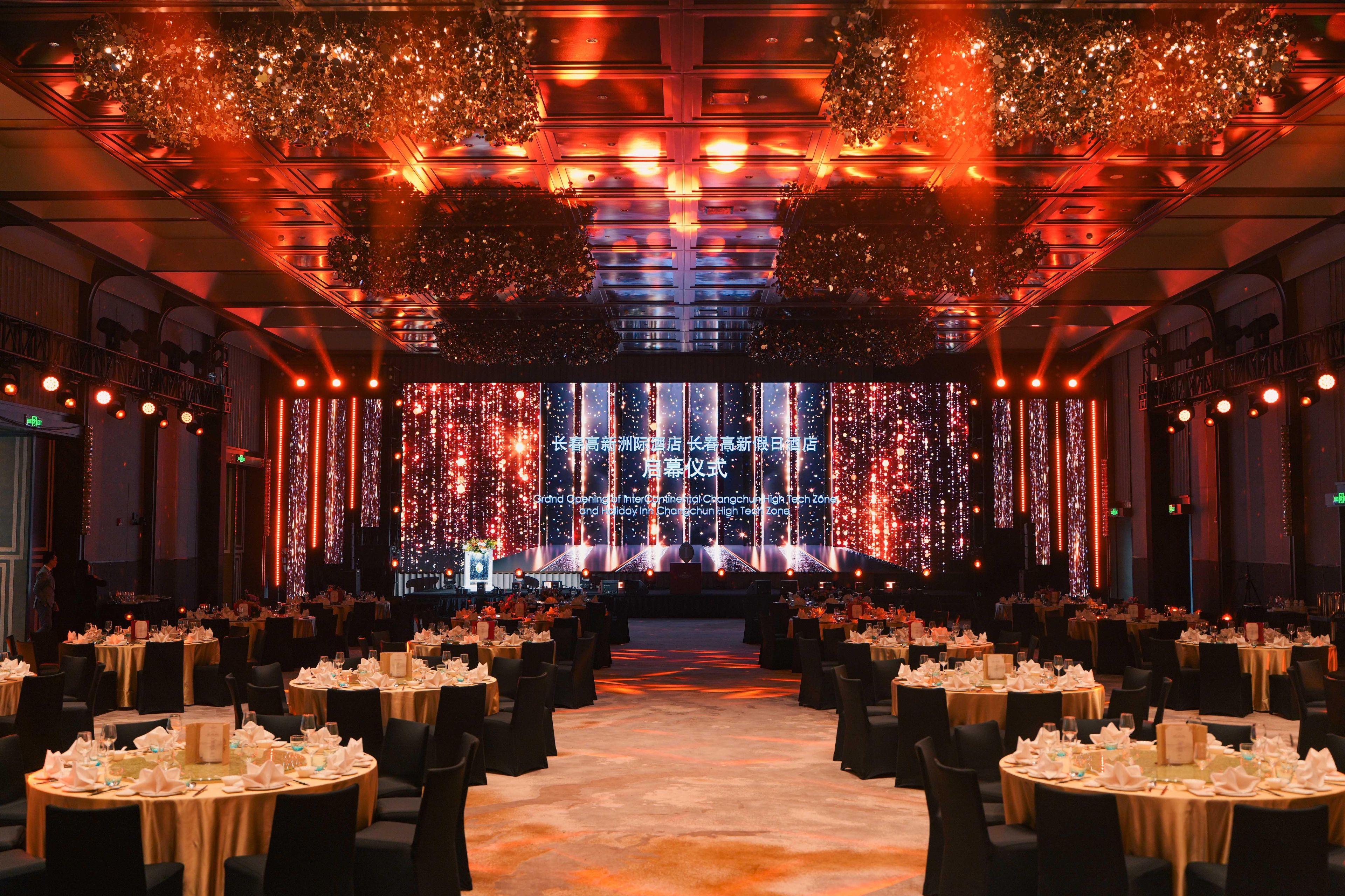Un salón de banquetes con mesas redondas dispuestas para un evento formal, que cuenta con un escenario, una pantalla grande e iluminación decorativa.