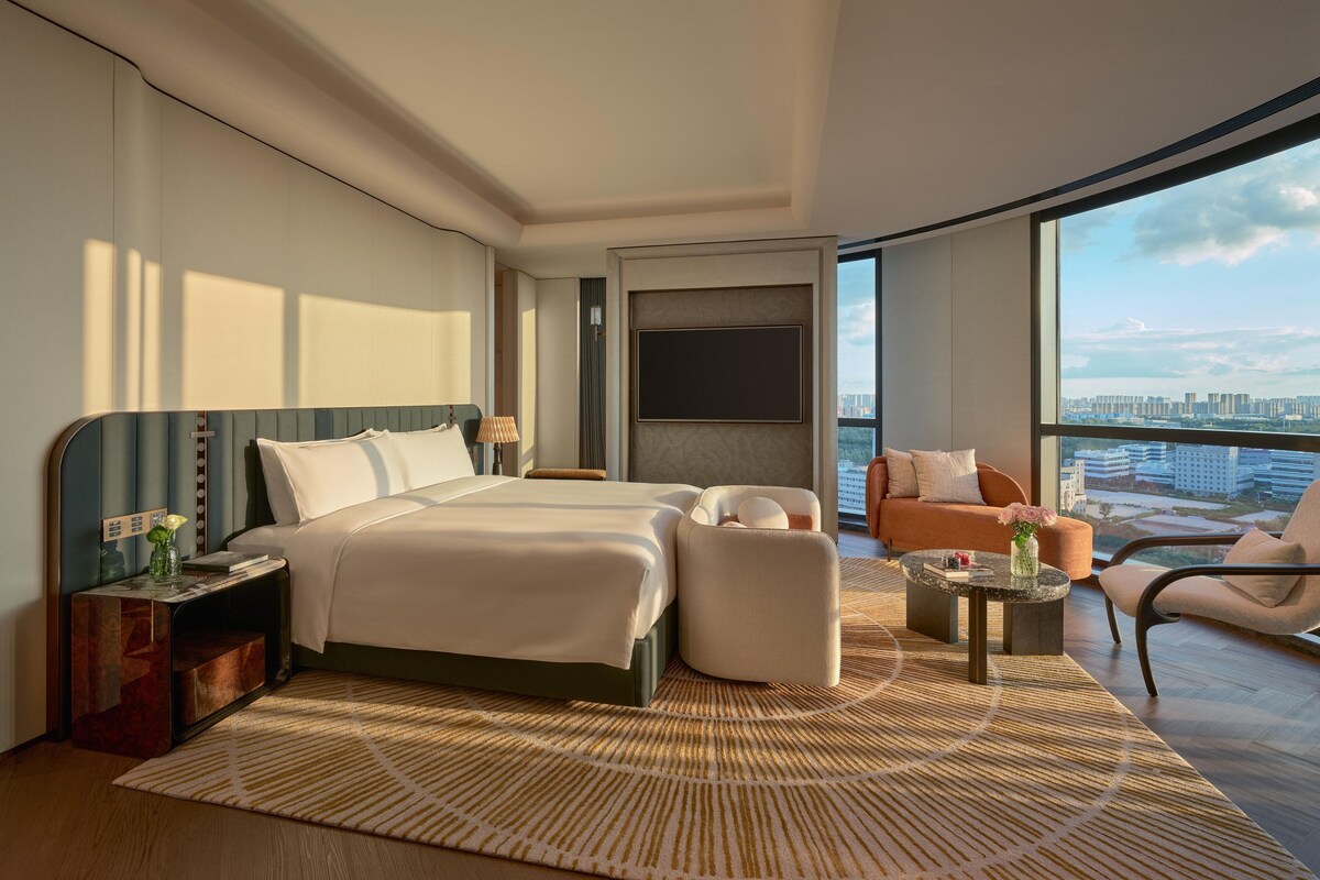 Een moderne hotelkamer met een groot bed, twee nachtkastjes, een zithoek met een bank en fauteuils, een klein bijzettafeltje en een groot raam met uitzicht op de stad.