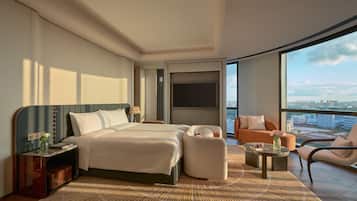 Una habitación de hotel moderna con una cama grande, dos mesitas de noche, un área de estar con un sofá y sillones, una pequeña mesa de centro y un ventanal con vistas a la ciudad.