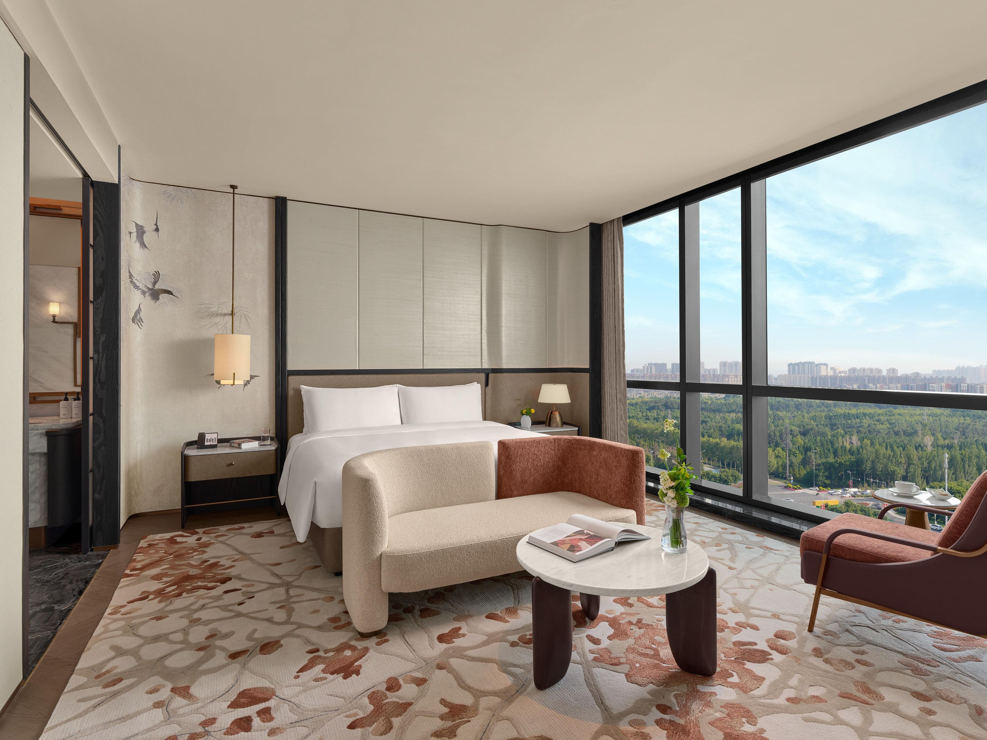 A hotel room with a bed, a sofa, a small table, and a view of a cityscape through large windows.