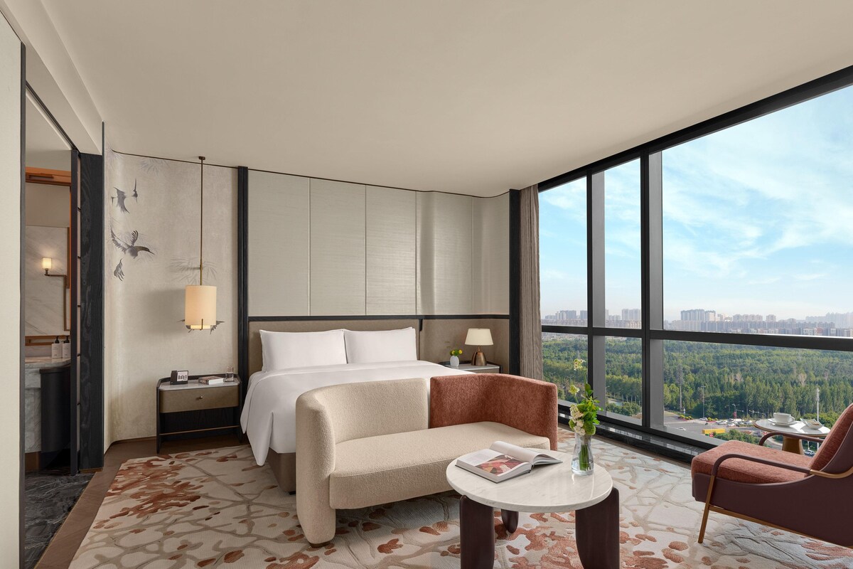 A hotel room with a bed, a sofa, a small table, and a view of a cityscape through large windows.