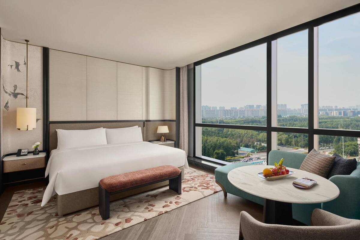 Een moderne hotelkamer met een groot bed, een bankje, een ronde tafel met fruit en uitzicht op een stadsbeeld door grote ramen.