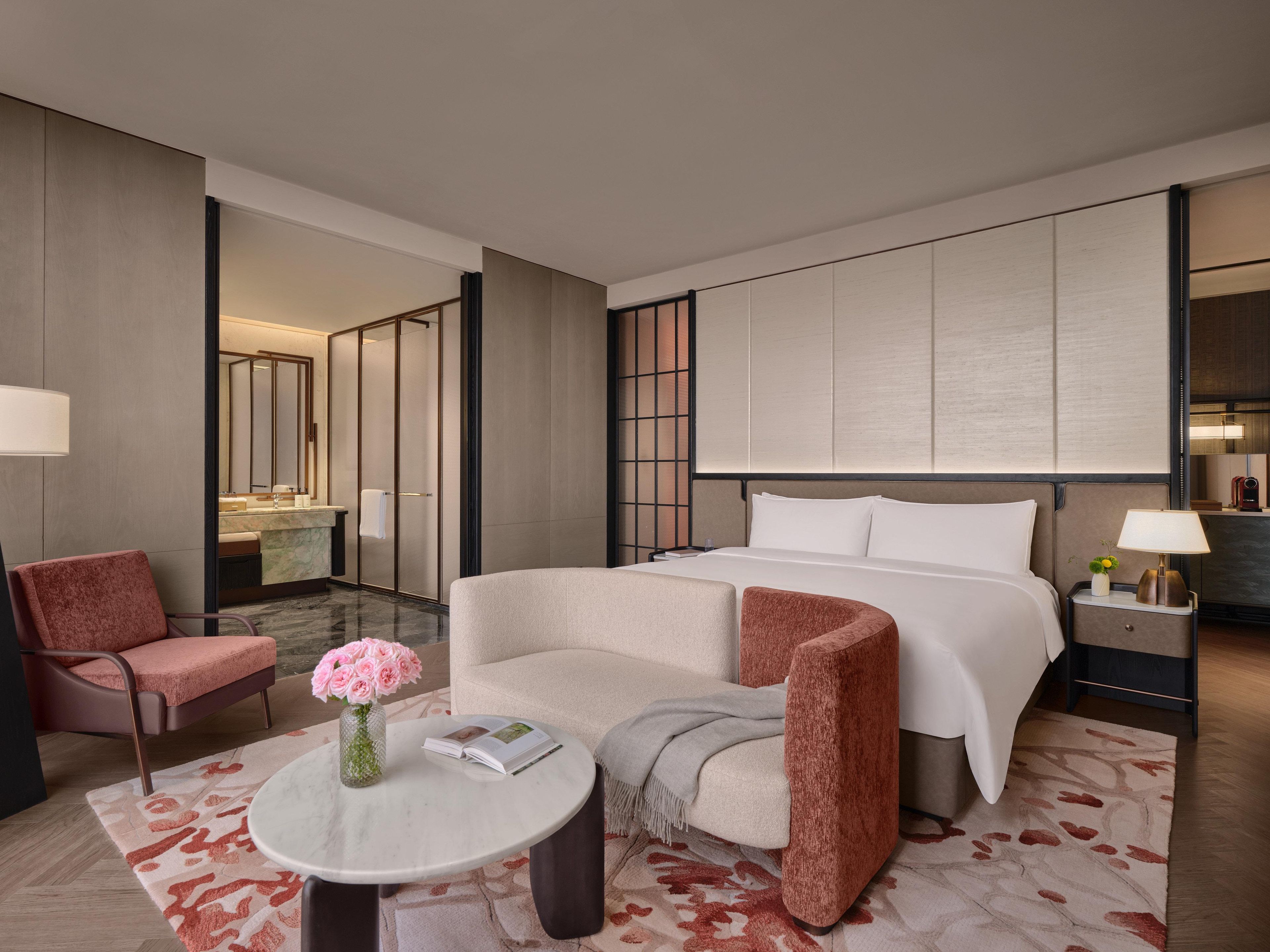 Une chambre d’hôtel comprenant un lit, deux fauteuils, une petite table avec un vase de fleurs, une table de chevet avec une lampe et un grand miroir au mur.