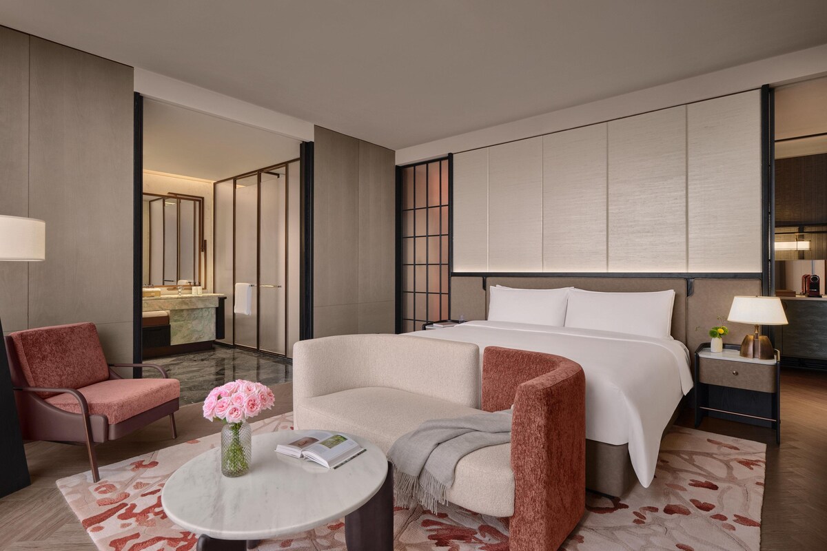 A hotel room with a bed, two armchairs, a small table with a vase of flowers, a nightstand with a lamp, and a large mirror on the wall.