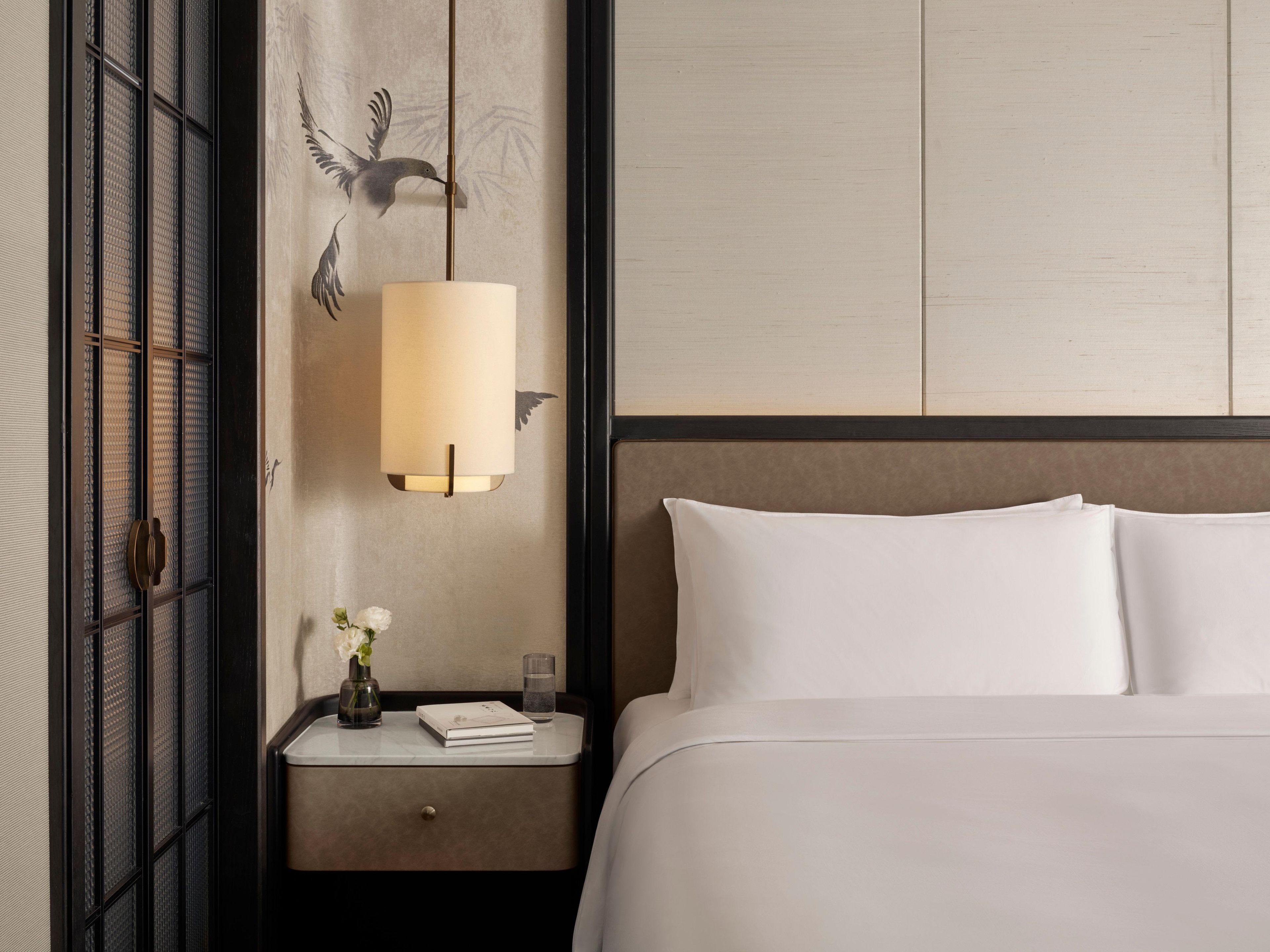 Een net opgemaakt bed met witte lakens, een nachtkastje met een lamp, een vaas met bloemen en een muur met een decoratieve vogelmuurschildering.