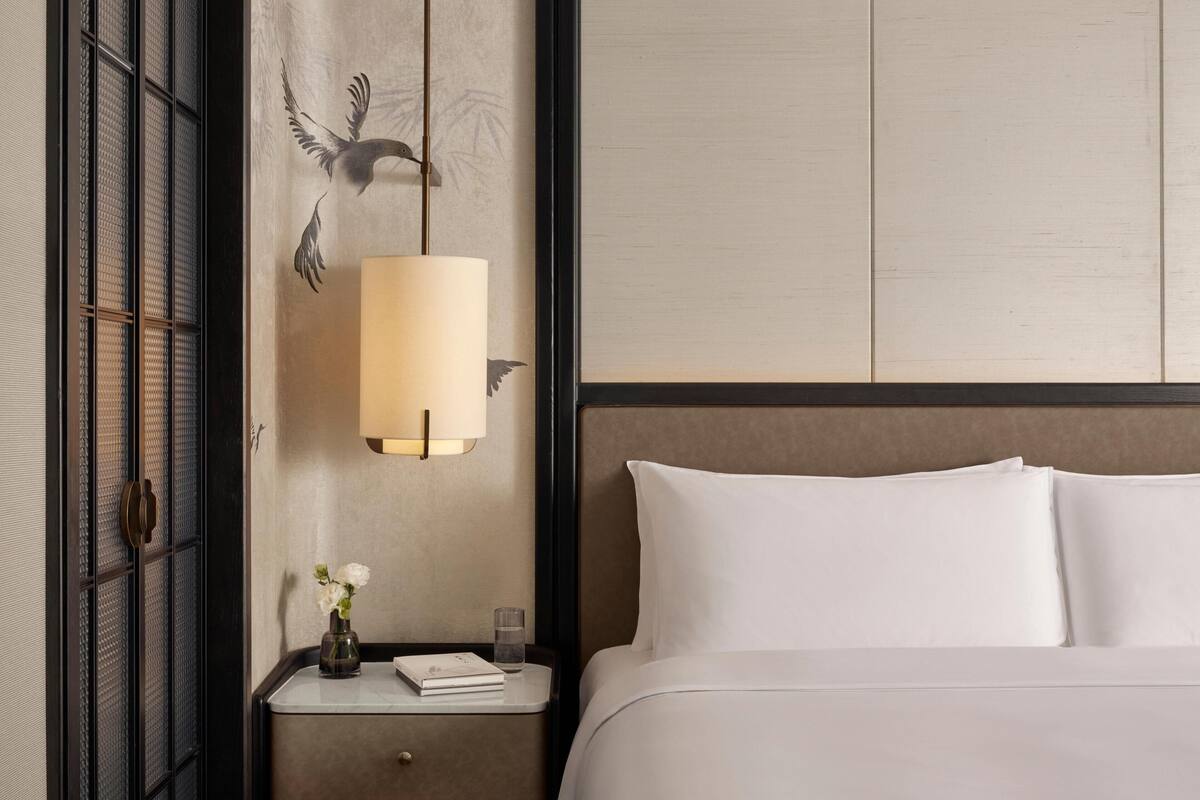 Een net opgemaakt bed met witte lakens, een nachtkastje met een lamp, een vaas met bloemen en een muur met een decoratieve vogelmuurschildering.